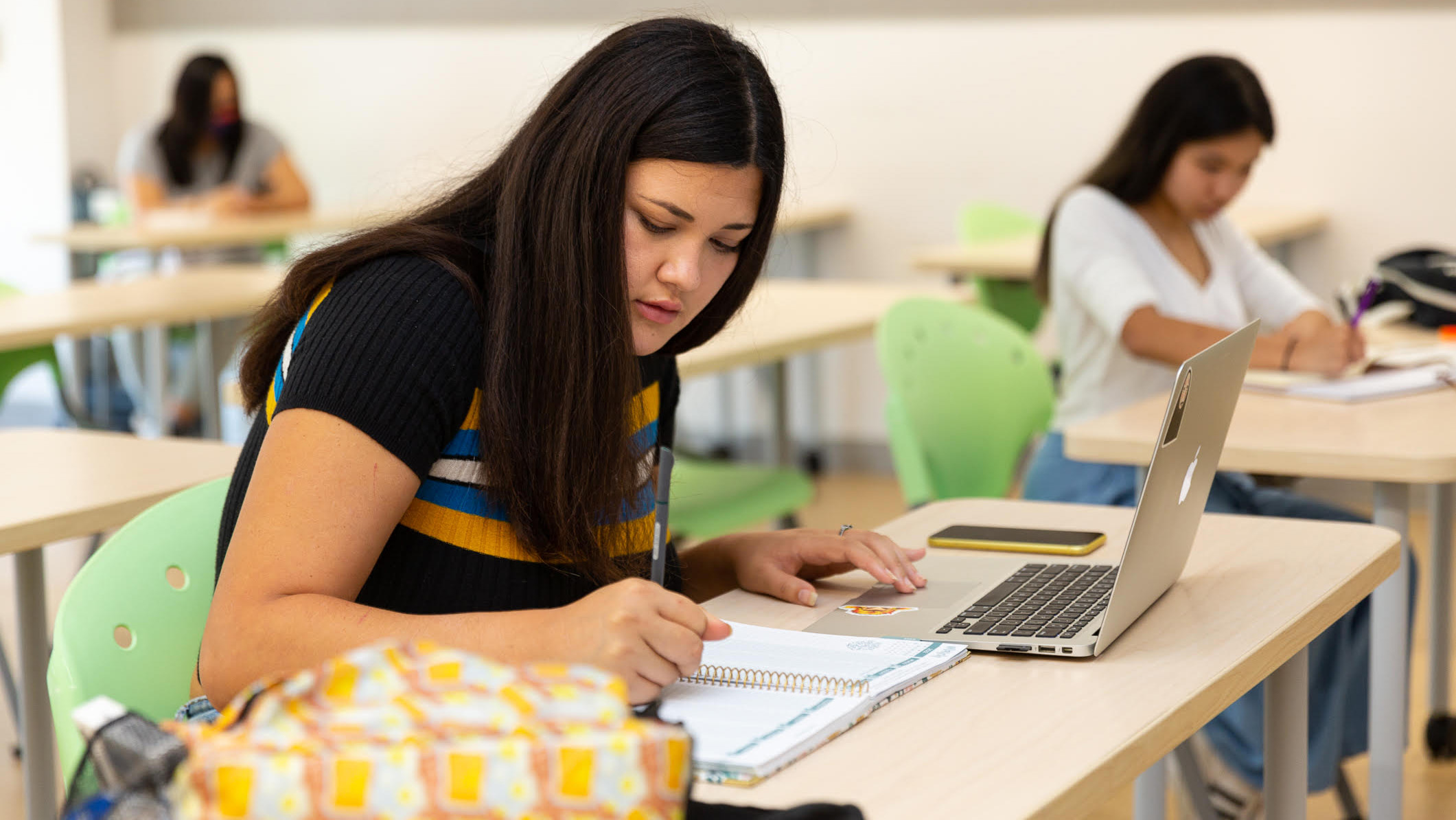 A student writing in a notebook with a laptop, with other students studying in the background.