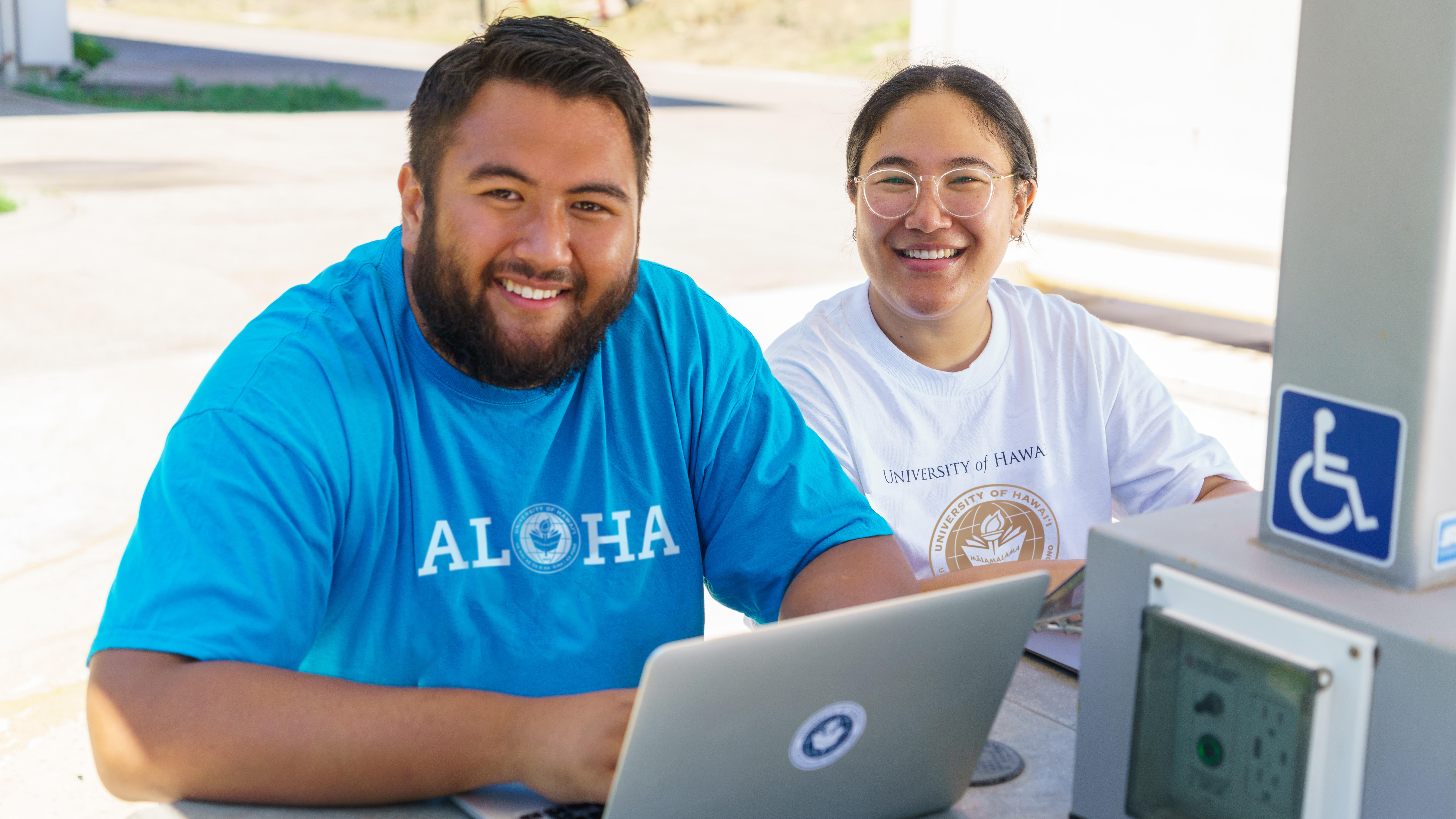 Two students smiling with laptops, sitting outside at a table.